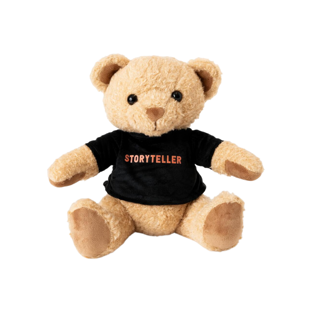 Light tan plush teddy bear wearing a black short-sleeve T-shirt with “Storyteller” printed across the chest in orange-red lettering, shown against a white background.