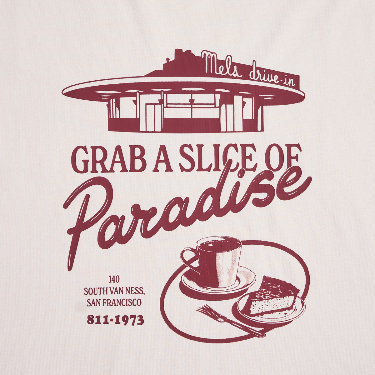 A close-up of a beige colored t-shirt featuring a vintage-style maroon graphic print of a retro diner with the text 'GRAB A SLICE OF Paradise' atop a coffee and cake illustration.