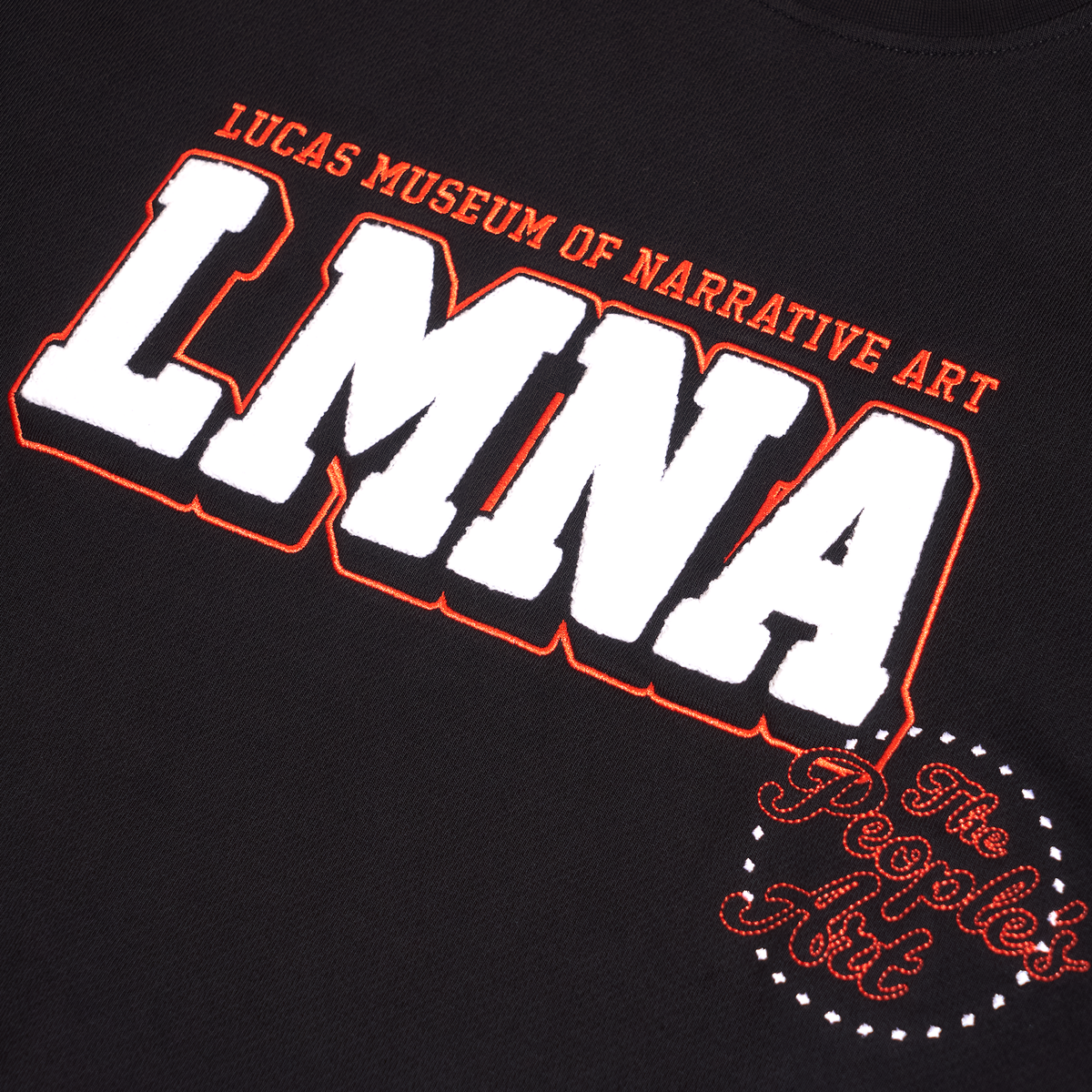 Close-up of a black graphic t-shirt featuring the text 'LUCAS MUSEUM OF NARRATIVE ART LMNA' in bold white letters with orange and black outlines, and a circular 'The People's Art' logo in orange and white.