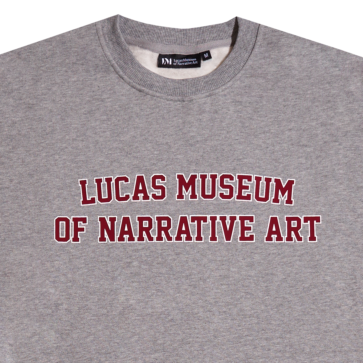 A close-up of a heather gray crewneck sweatshirt featuring the text 'LUCAS MUSEUM OF NARRATIVE ART' printed in red varsity-style letters with a white outline across the chest.