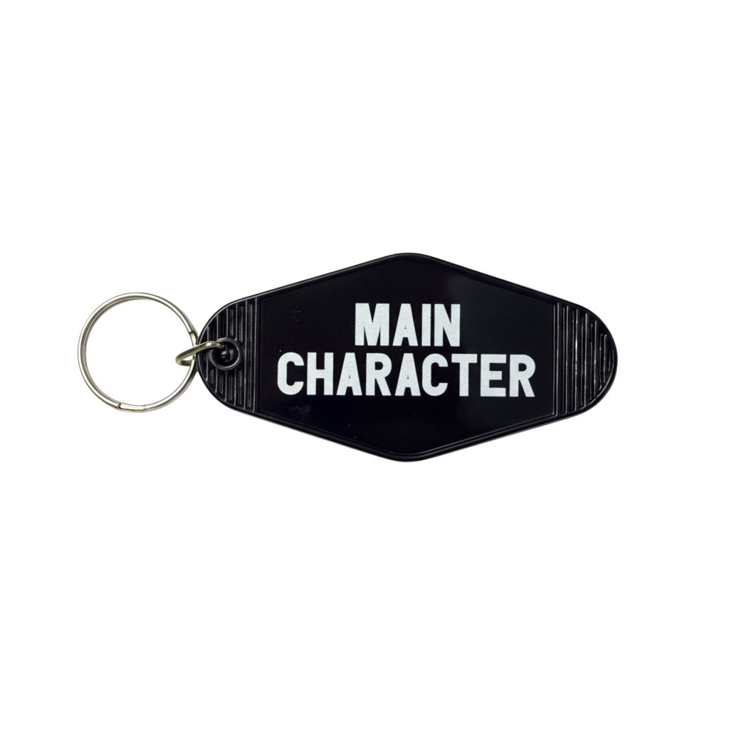 A black plastic keychain with a silver metal split ring, featuring the words 'MAIN CHARACTER' printed in bold white capital letters on a black background.