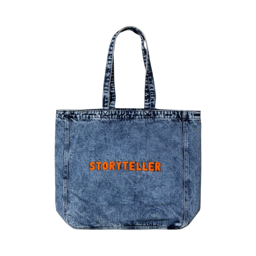 A blue acid-wash denim tote bag with 'STORYTELLER' printed in orange across the front, set against a plain white background.
