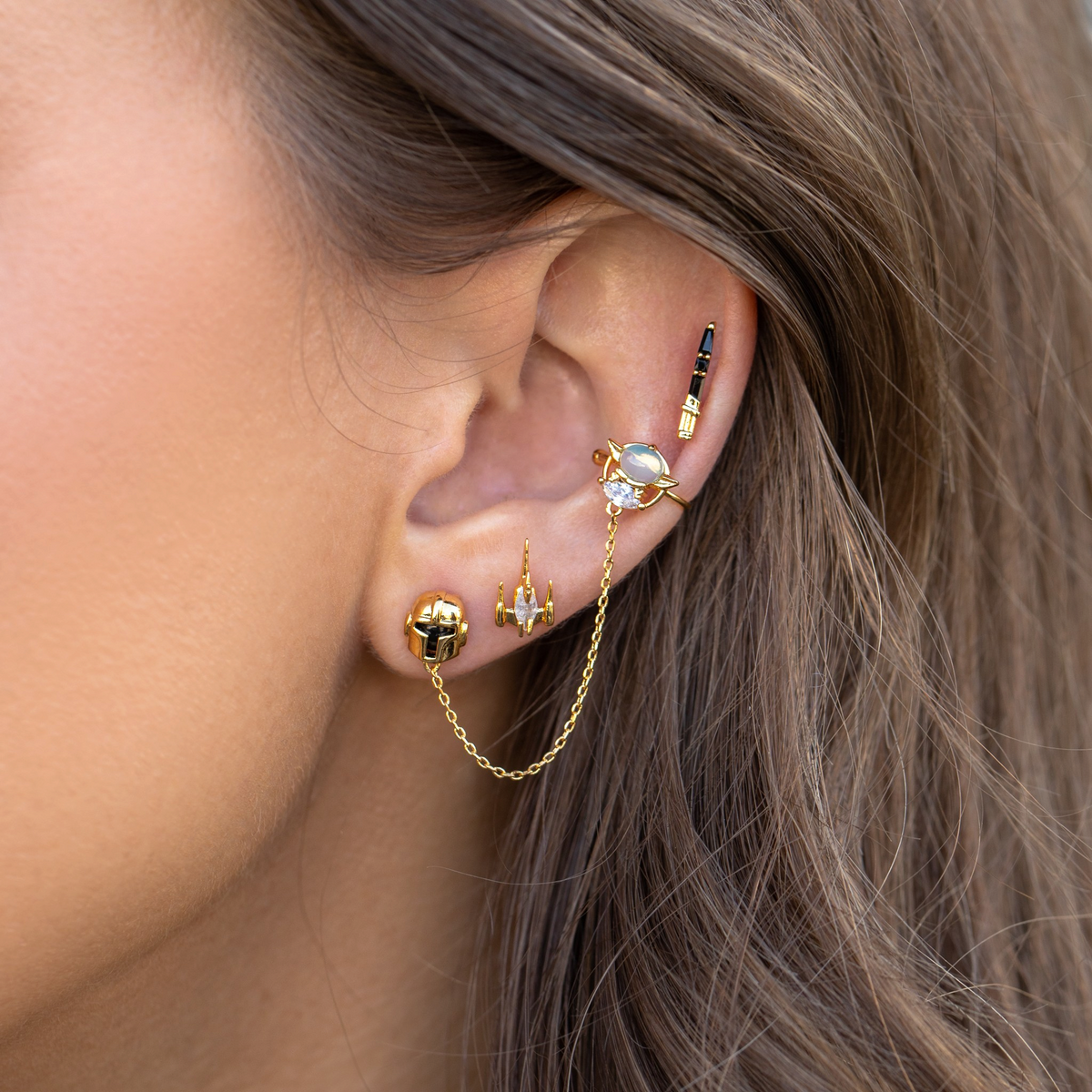 Four-piece gold-tone stud earring set featuring a helmet-inspired stud with chain detail, a starfighter motif, a lightsaber-inspired stud, and a celestial design with an iridescent cabochon and clear crystal accent, photographed on a model's ear.