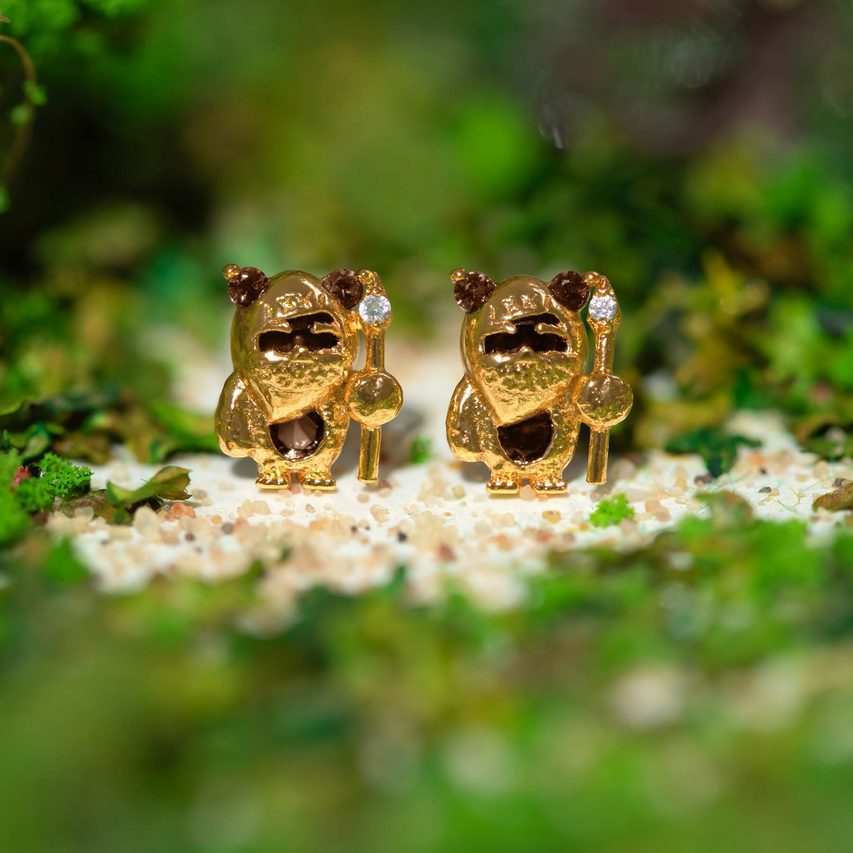 Gold-tone Ewok stud earrings featuring sculpted character designs with brown crystal accents and staff details, photographed on a bed of white sand with a blurred green mossy background.