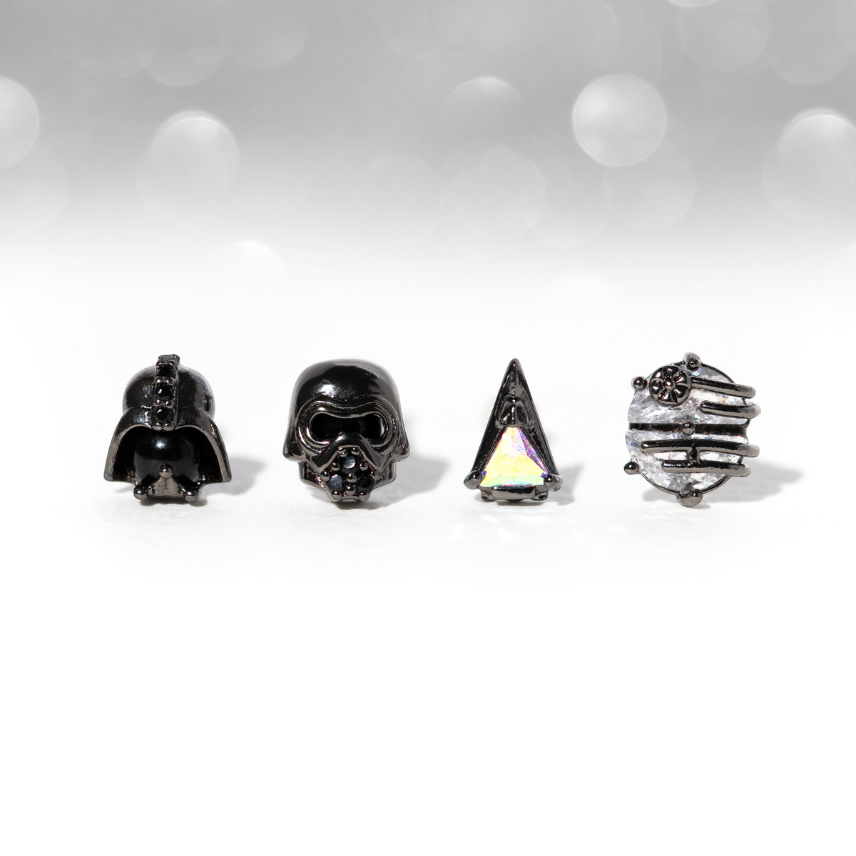 Four-piece gunmetal-tone stud earring set featuring villain-inspired helmet motifs, a starship design, and an iridescent triangular crystal stud, shown on a white surface against a soft gray bokeh background.