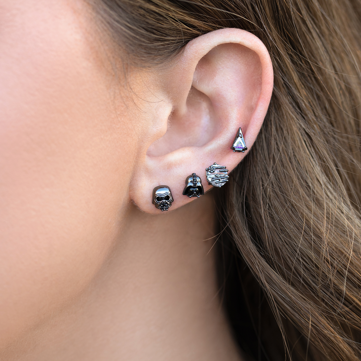 Four-piece gunmetal-tone stud earring set featuring villain-inspired helmet motifs, a starship design, and an iridescent triangular crystal stud, photographed on a model's ear.