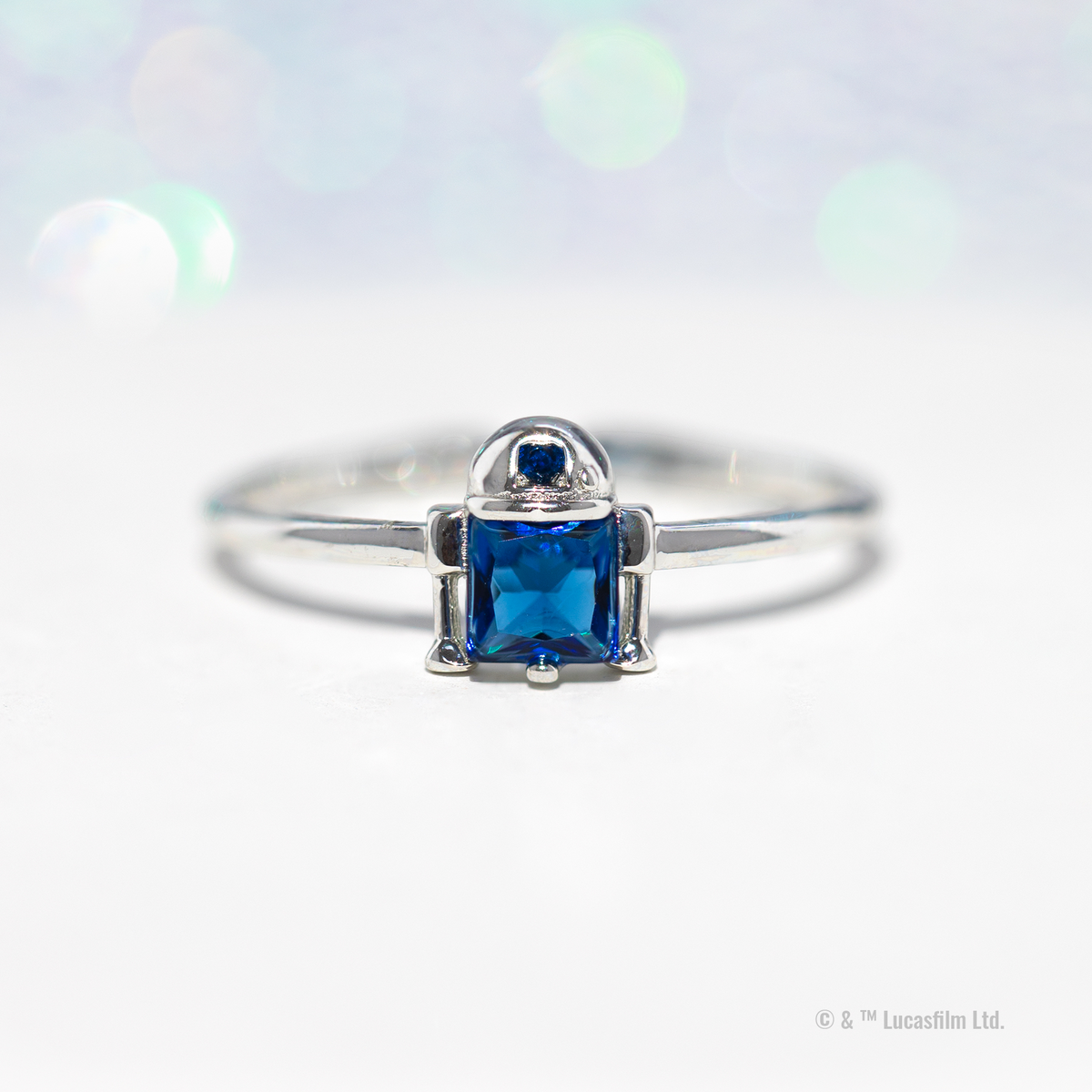 Silver-tone R2-D2 ring featuring a stylized droid-inspired design with a blue crystal centerpiece and blue accent stone, photographed against a bright white background with soft green and blue bokeh lights.