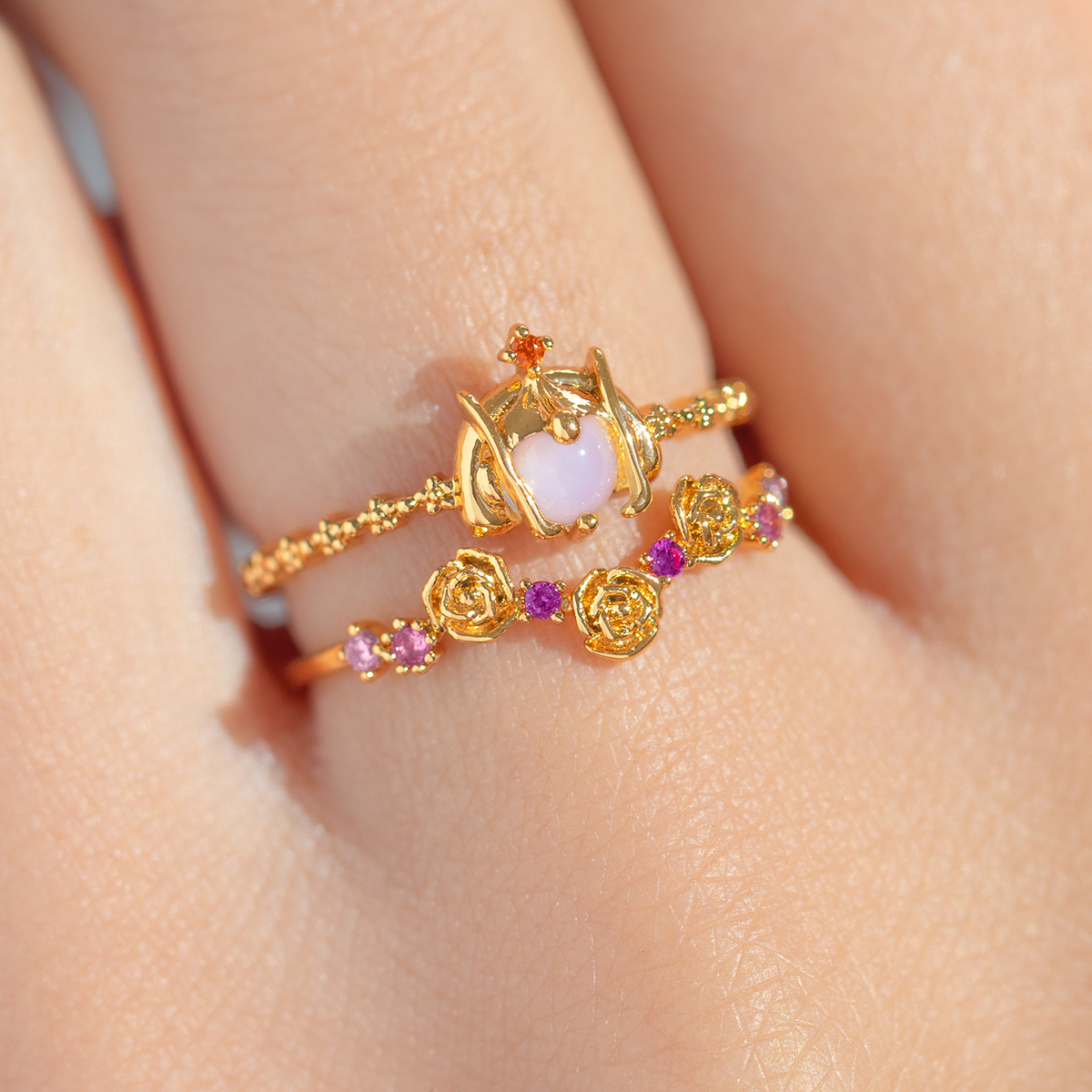 Two-piece gold-tone Padmé Amidala ring set featuring an ornate crown motif with a white stone and a matching rose-pattern band with pink gemstone accents, worn on a model's hand.