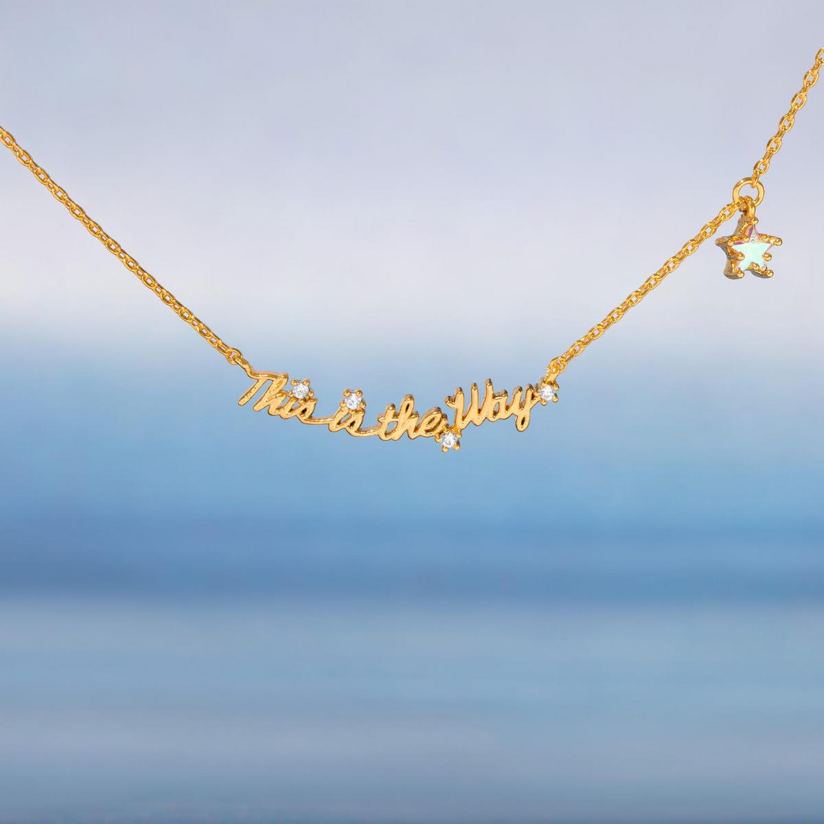 Gold-tone necklace featuring the phrase “This Is the Way” in script lettering, accented with clear crystals and a dangling iridescent star charm, shown on a gradient blue background.