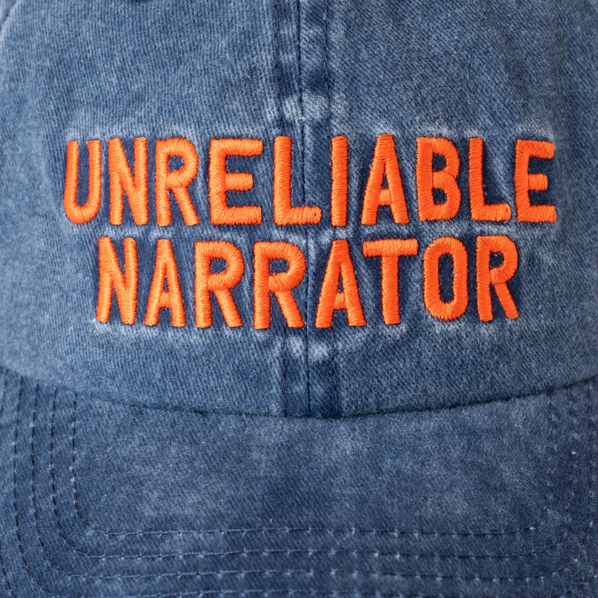 Washed cotton cap featuring embroidered text reading “Unreliable Narrator,” with an adjustable back strap and metal fastener. Interior seams are finished with logo-branded binding.